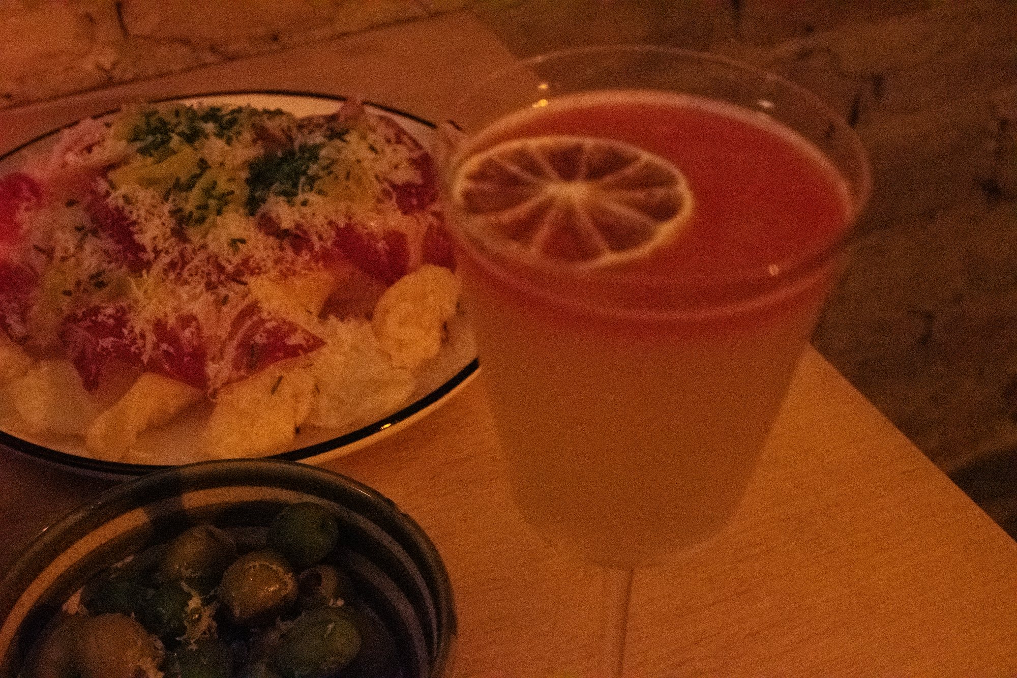 A plate of loaded chips with herbs and parmesan, a bowl of marinated olives, and an orange cocktail with a citrus-wheel garnish, lit by candlelight