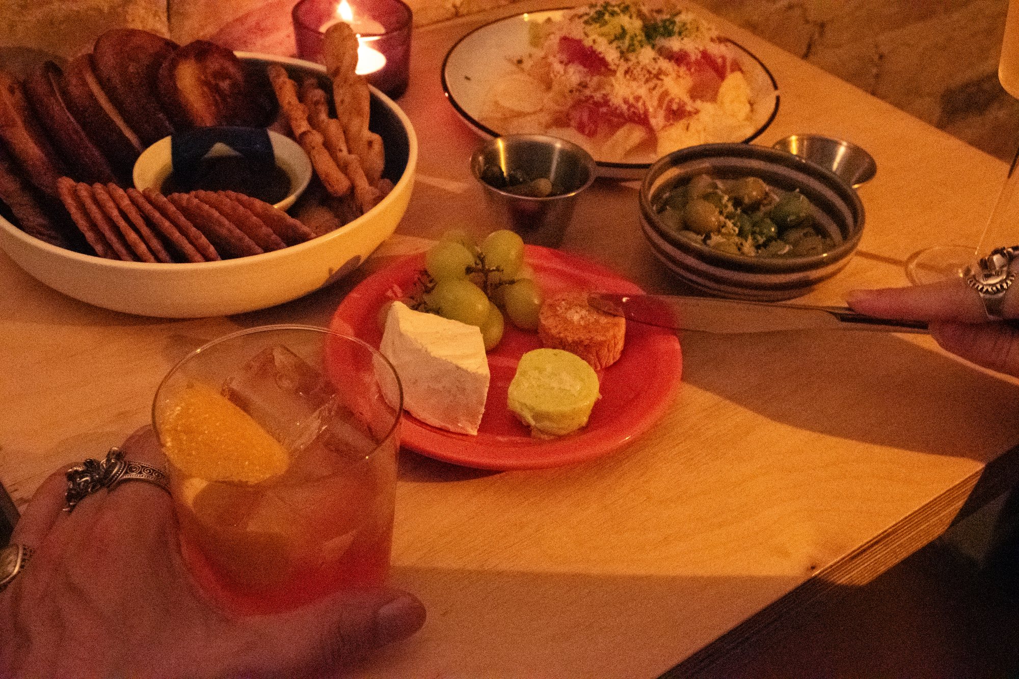 A spread of crackers, breadsticks, cheese, and grapes beside a held rocks-glass cocktail with an orange twist